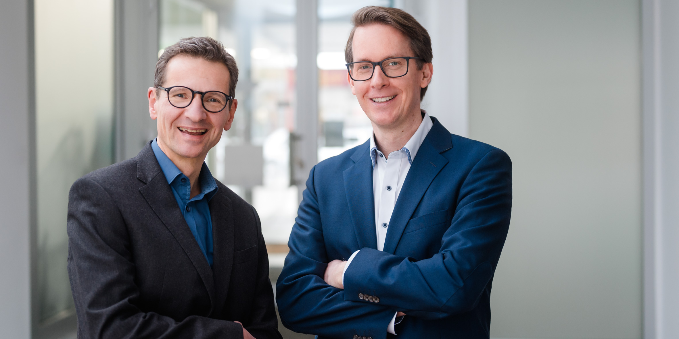 Die Partner Gottfried Müller (links, mit runder Brille und dunklem Sakko) und Anton Mooseder (rechts, im blauen Sakko) lächelnd in einem hellen Büro