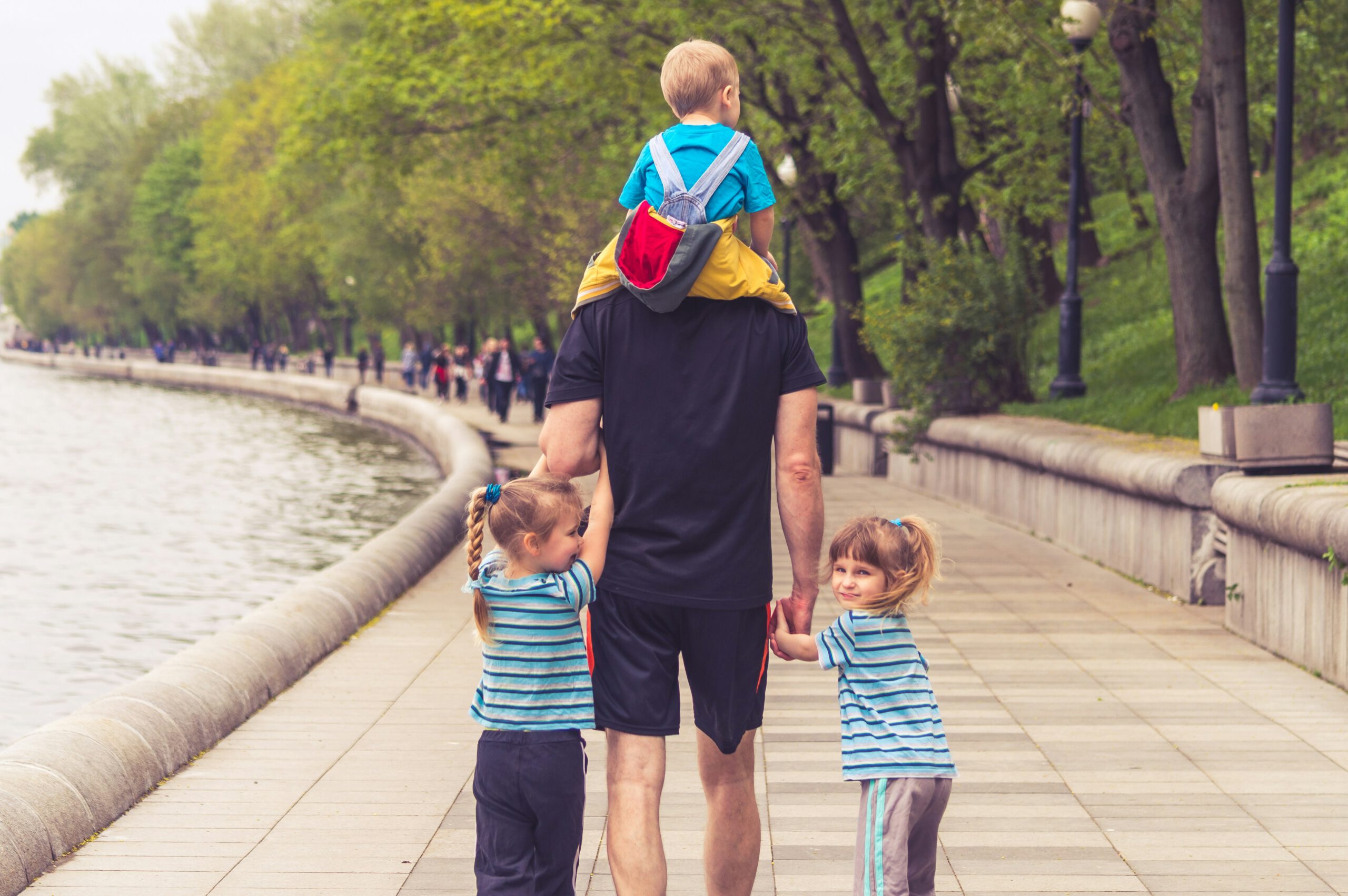Ein Vater geht mit drei kleinen Kindern an einer Uferpromenade spazieren, ein Kind auf den Schultern, zwei an der Hand – Sinnbild für Zeit mit der Familie dank attraktiver Dienstplanung